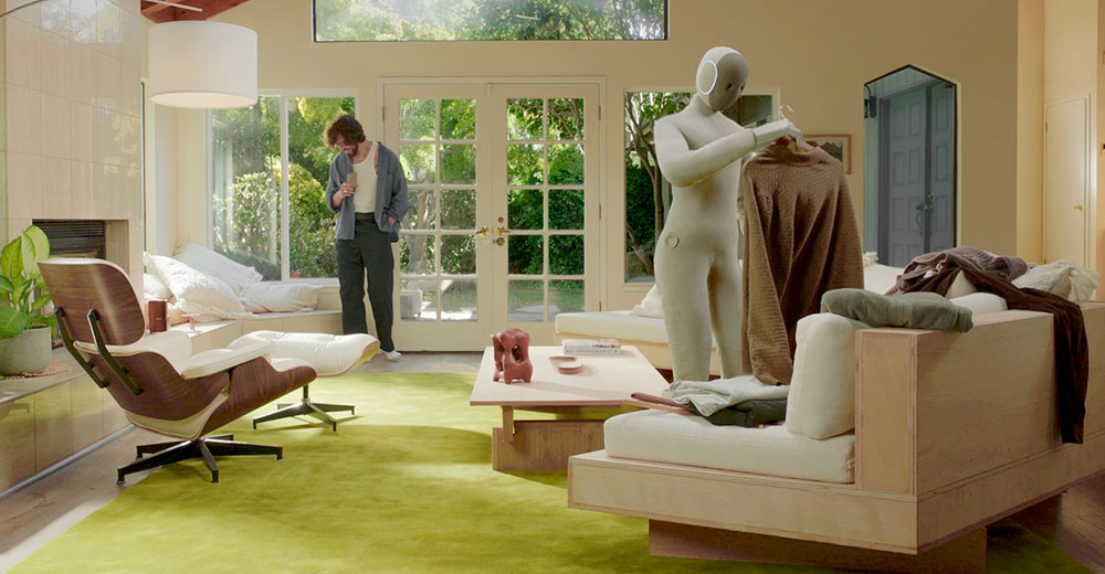 Humanoid home robot folding clothes in a modern living room while a person stands nearby