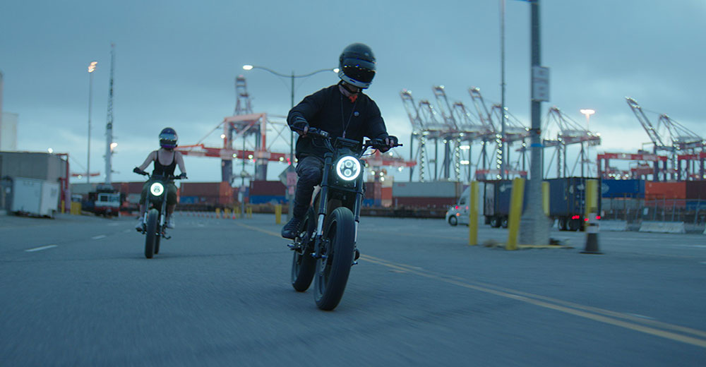 Two riders on Onyx RCR electric motorbikes traveling through an industrial port area